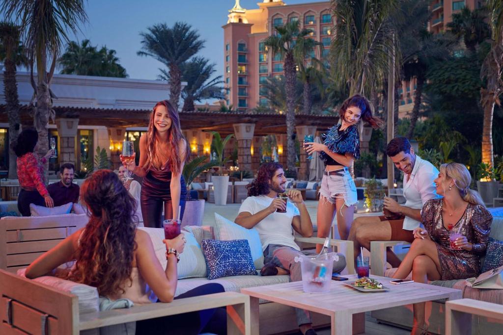 Vakarā izklaidējoša atmosfēra pie baseina, kur jaunieši baudīja kokteiļus un sarunas, izceļot draudzību un jautrību. Eleganti tērpi un tropiskā vide rada ideālu fonu brīvdienu noskaņai.