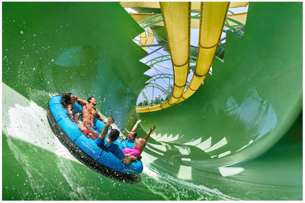 Grupas cilvēku jautrā braucienā ar riepu pa ūdens slidkalniņu, izbaudot adrenalīnu un izsistot ūdeni. Vivid zaļš fons un dzeltenas caurules papildina aizraujošo pieredzi.