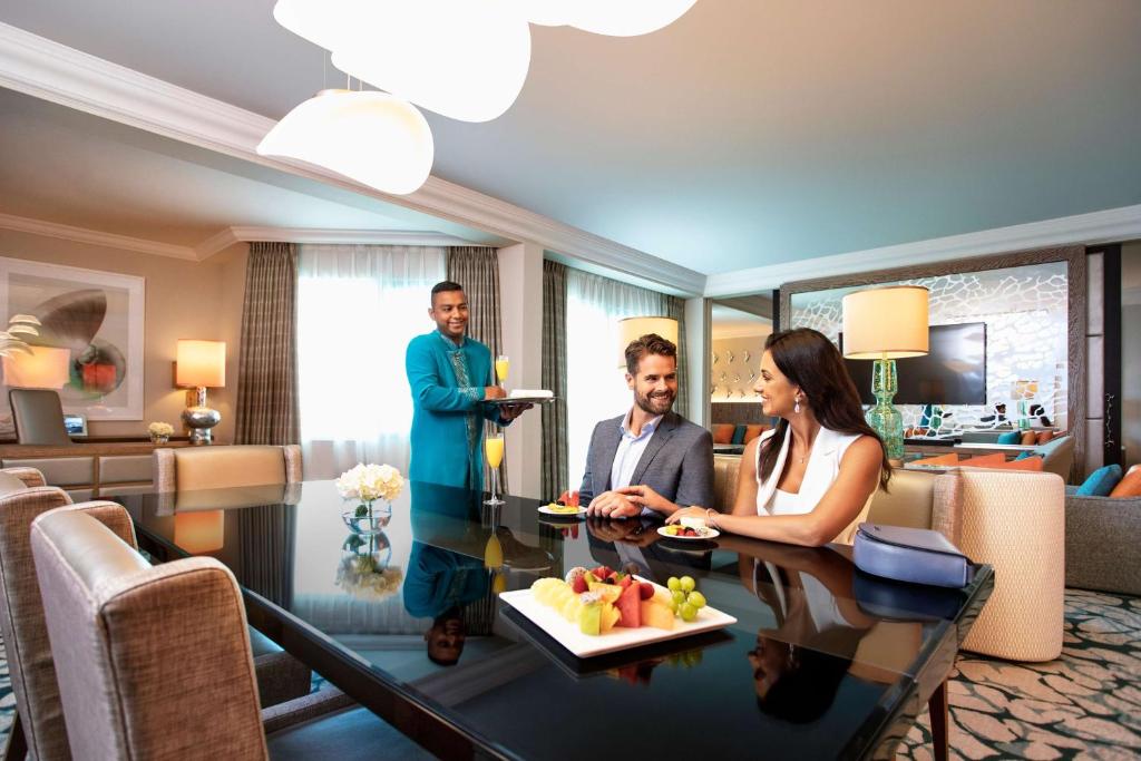 Luksusa viesnīcas numurs ar pāru romantisku brīdi, kur viesmīlis pasniedz ēdienu un dzērienus pie elegantā galda.