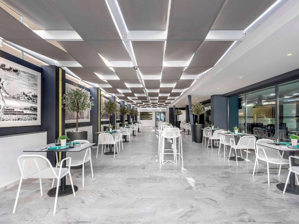 Mūsdienīgs restorāns ar elegantu interjeru, baltiem galdiem un krēsliem, kā arī zaļiem augiem dekorācijā. Gaismas un arhitektūras elementi rada patīkamu atmosfēru.