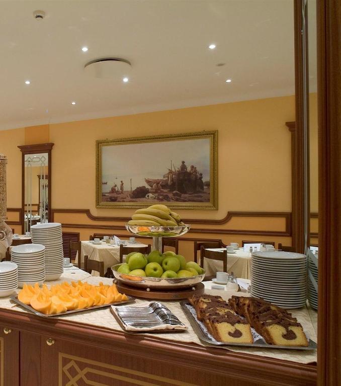 Restorāna interjers ar augļu un konditorejas izstrādājumu galdu, uz kura redzami banāni, zaļie āboli un kūkas. Fons rotā glezna ar jūras ainavu.