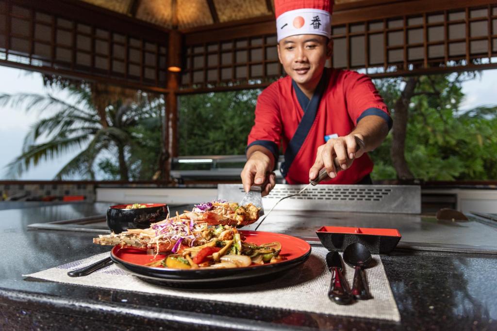 Šefpavārs, tērpies sarkanā apģērbā, pasniedz gardus ēdienus uz galda, kam blakus atrodas gatavošanas piederumi un trauki. Aiz viņa redzama tropu ainava ar palmām.