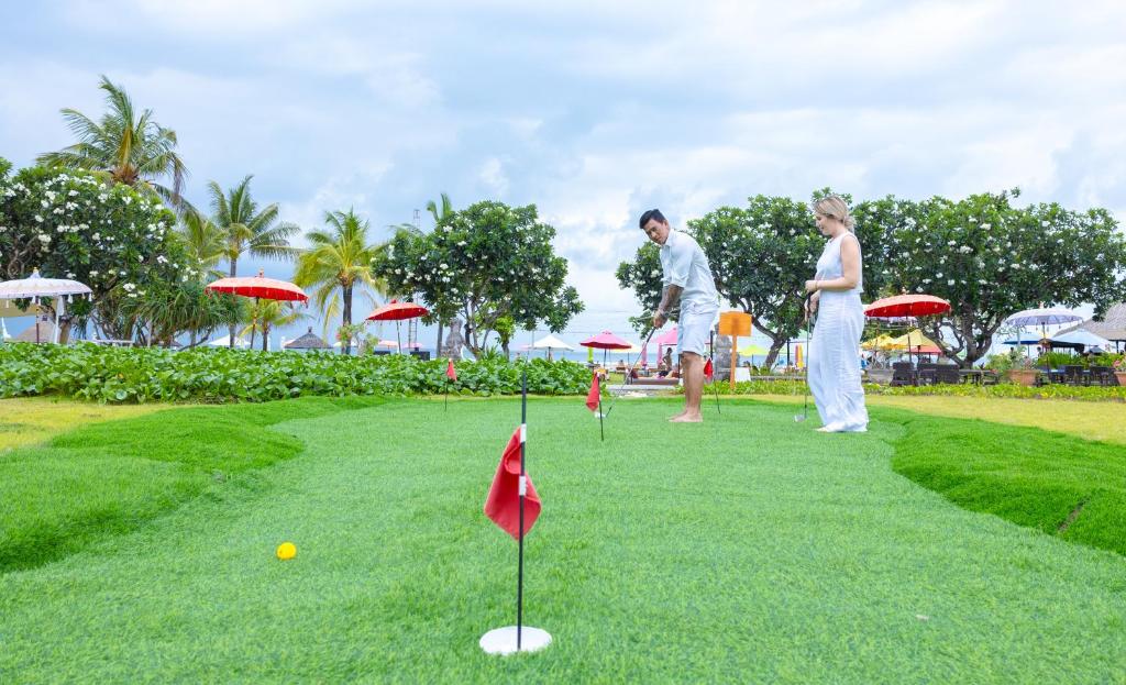 Divi cilvēki spēlē mini golfu tropiskā vidē ar palmām un saules lietussargiem. Zāle ir zaļa un labi kopta, radot relaksējošu atmosfēru.