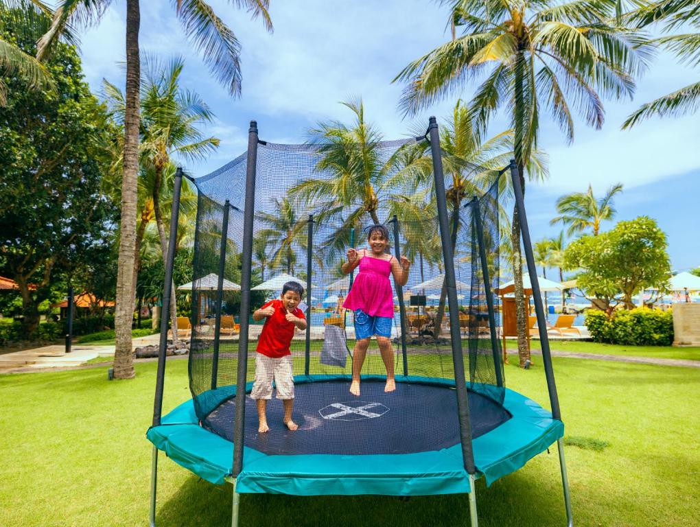 Bērni lecot uz trampolīna, apkārt augot palmu kokiem un zaļai zālītei, radot priecīgu un izklaidējošu atmosfēru.