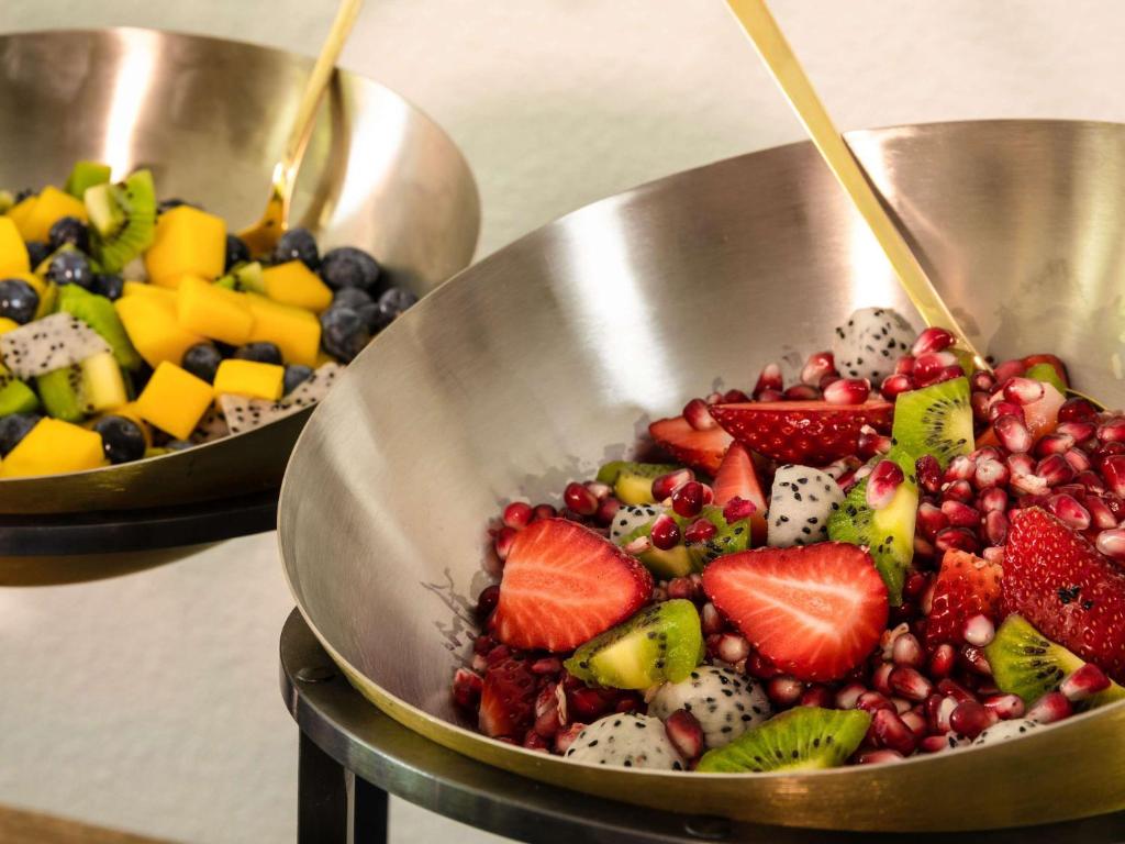 Divas augļu salātu bļodas, viena ar mango, kiwi un mellenēm, otra ar zemenēm, granātāboliem un pūķa augļiem, pasniegtas modernā traukā.