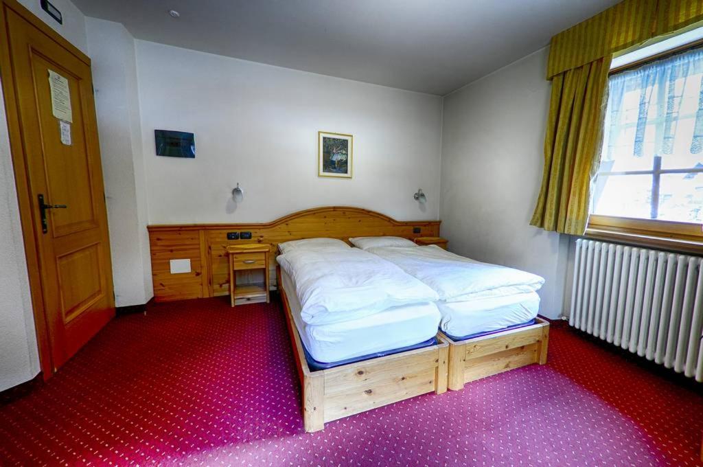 Mājīgs viesnīcas numurs ar divām gultām, koka mēbelēm un sarkanu paklāju. Telpā ir logs ar dzeltenām aizkariem, kas nodrošina dabīgu apgaismojumu.