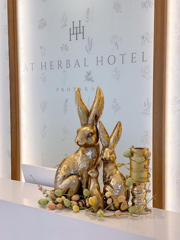 Zelta zaķi dekorē viesnīcas reģistratūru, papildināti ar krāsainām olām un ziediem. Viesnīcas nosaukums "AT HERBAL HOTEL" ir redzams fona paneļa augšdaļā.