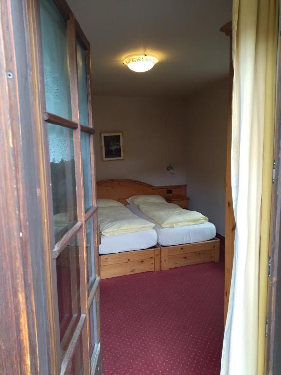 Gultas istaba ar divām atsevišķām gultām, redzama caur durvīm, ar koka mēbelēm un siltu apgaismojumu.