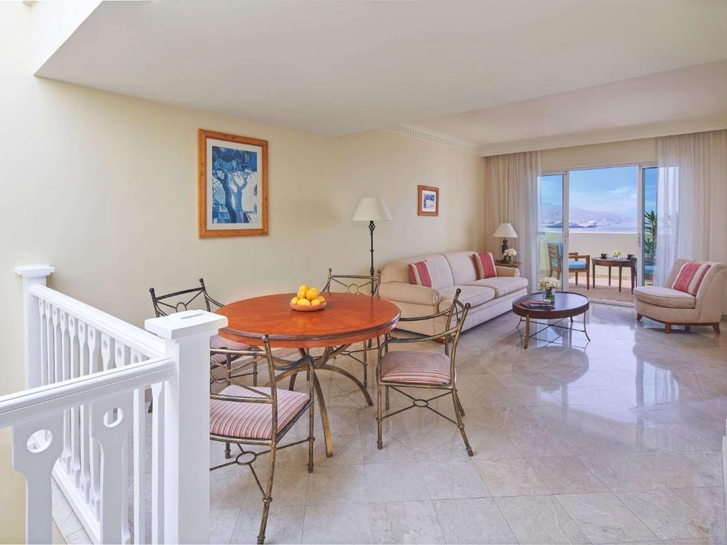 Gaiša viesistaba ar dīvānu, ēdamgaldu un skatu uz ārpusi, kur redzami kalni un dabiski elementi. Interjers ir elegants ar mūsdienīgu dizainu.