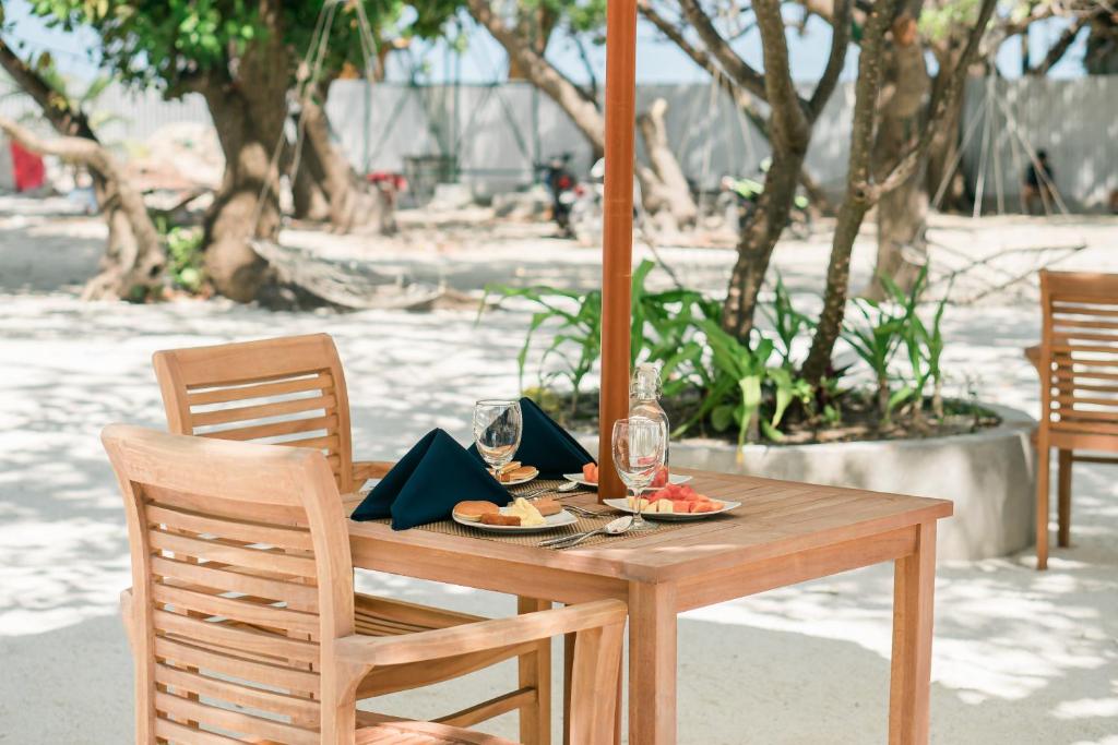 Koka galds ar divām krēslām ir izvietots zem nojumes, blakus zaļiem augiem un pludmales smiltīm. Uz galda ir trauki ar augļiem un dzeramais ūdens, radot relaksējošu atmosfēru.
