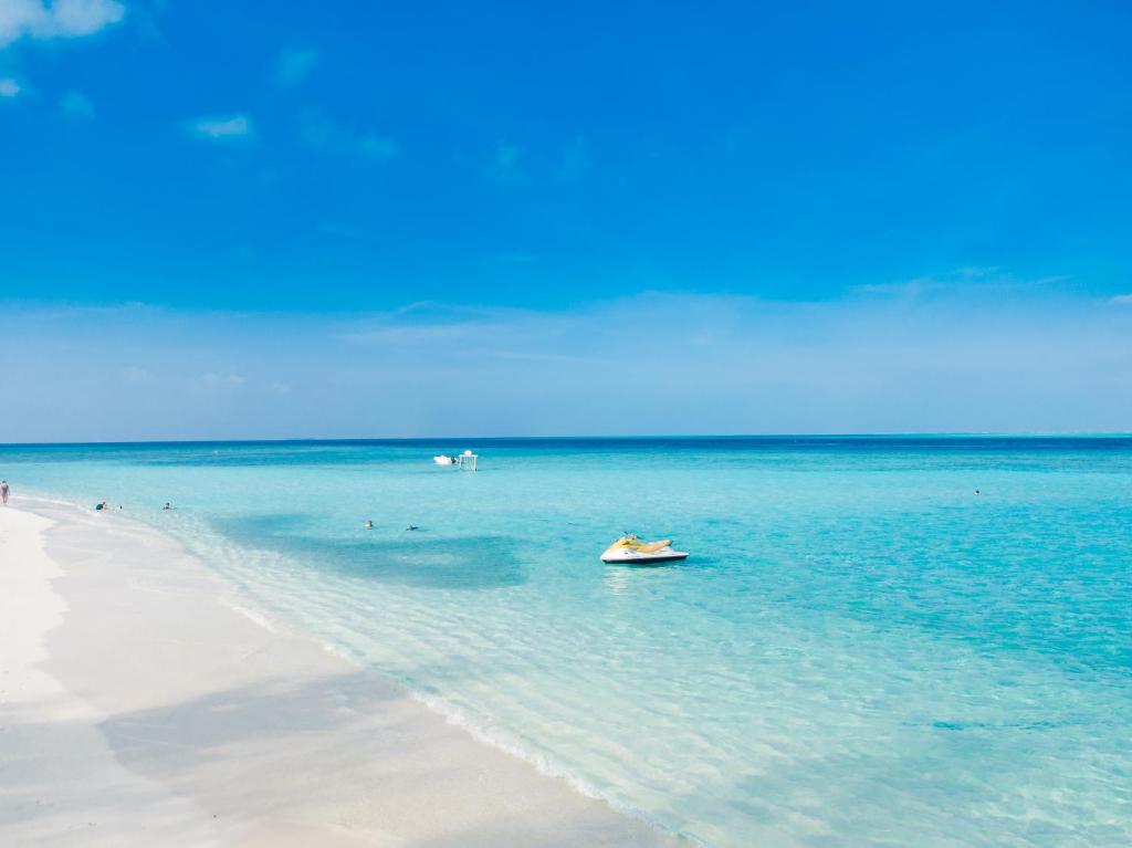 Skaists pludmales skats ar dzidru zilo ūdeni un baltu smiltīm, kur cilvēki bauda sauli un peldēšanu. Peld laiva pie krasta papildina mierīgu ainavu.