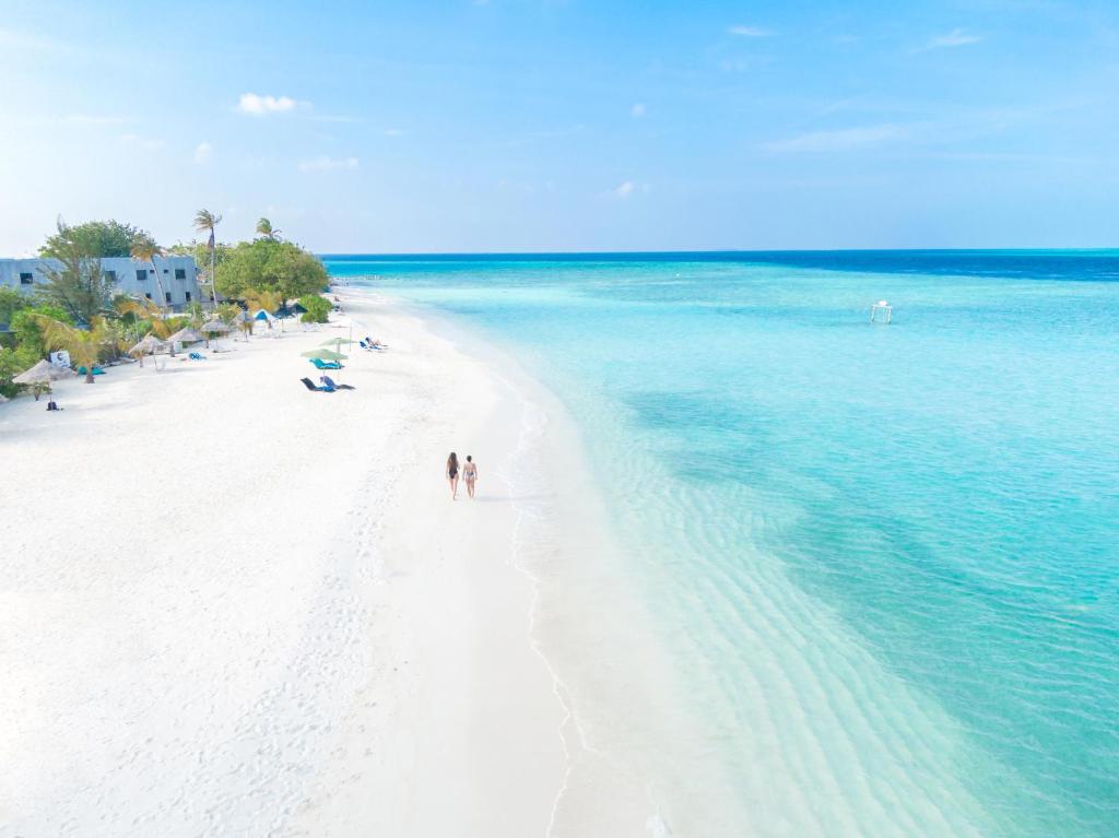 Divas personas staigā pa baltu pludmali, apņemta ar kristāldzidru ūdeni un palmām. Aiz viņām redzami sauļošanās krēsli un pludmales ēkas.