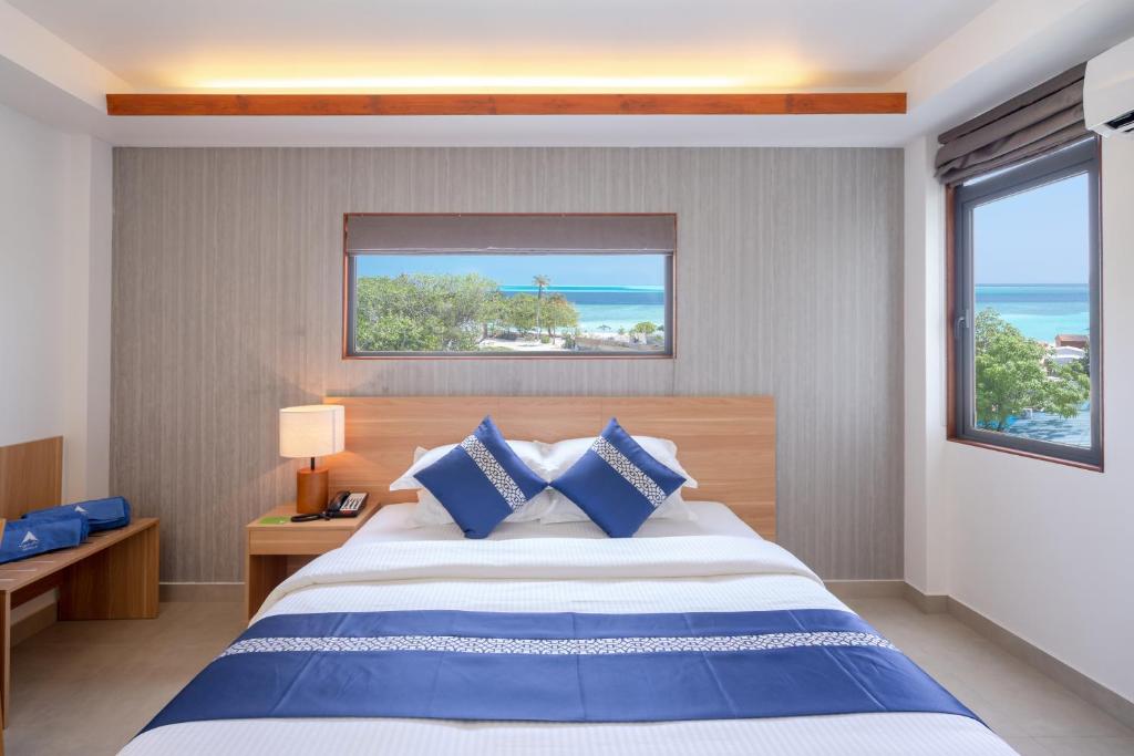 Mūsdienīgs guļamistabas interjers ar skatu uz jūru, ietver ērtu gultu ar zilu spilvenu un koka mēbelēm. Logi ļauj ieplūst dabiskai gaismai un piedāvā gleznainu ainavu.