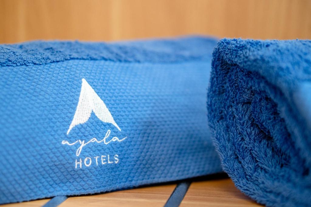 Zilā dvielī ar "Ayala Hotels" logotipu, kas uzsvērts ar balto kalnu simboliku, radot elegantu un viesmīlīgu atmosfēru.