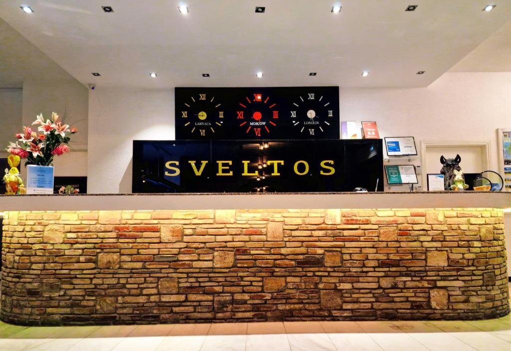 Recepcija viesnīcā "Sveltos" ar modernu dizainu, akmens apdari un pulksteņiem no dažādām pilsētām. Nozīmīgas detaļas ietver ziedus un informācijas stendus.