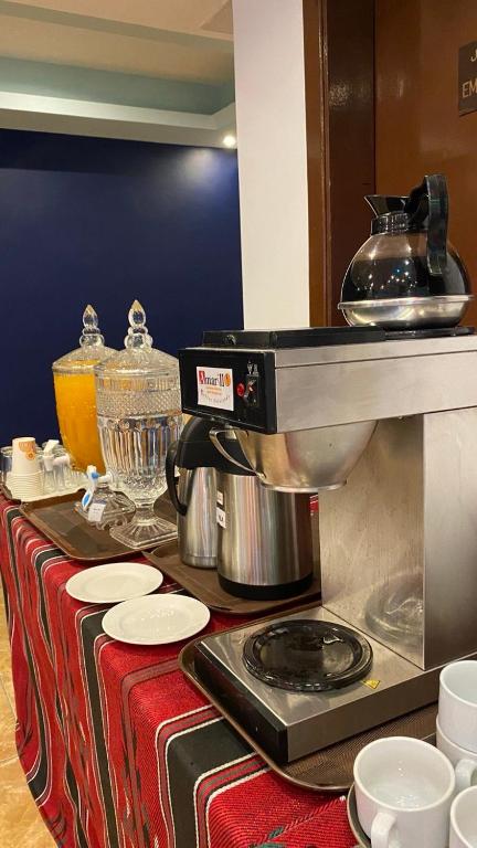 Kafijas automāts un dzērienu trauki uz galda, kurā redzami dažādi karstie un aukstie dzērieni, kā arī trauki un krūzes.