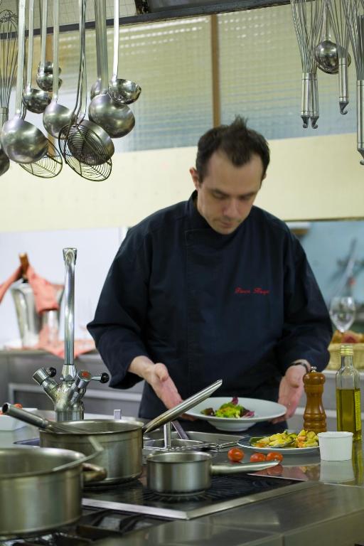 Šefpavārs gatavo ēdienu virtuvē, izmantojot dažādus virtuves rīkus un sastāvdaļas. Uz galda redzama šefpavāra pasniegta porcija ar dārzeņiem.