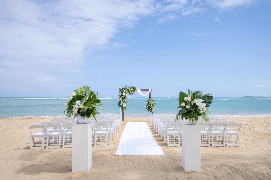 Pludmales kāzu ceremonijas vieta ar baltu paklāju, ziedu dekorācijām un skatu uz jūru. Rindas ar baltām krēslām gaida viesus svinībām.