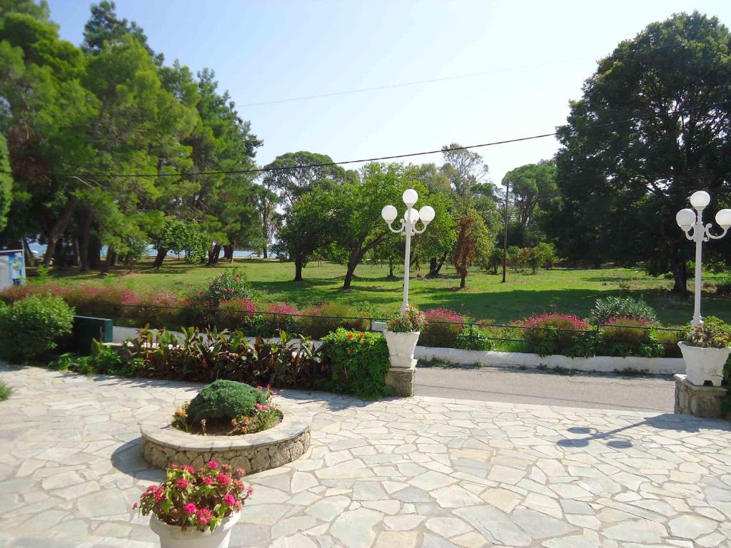 Zaļš parks ar koku rindām un ziedu dobēm, redzama akmens terase ar puķu podiem un laternām. Gaisīgs un saulains skats uz dabu.