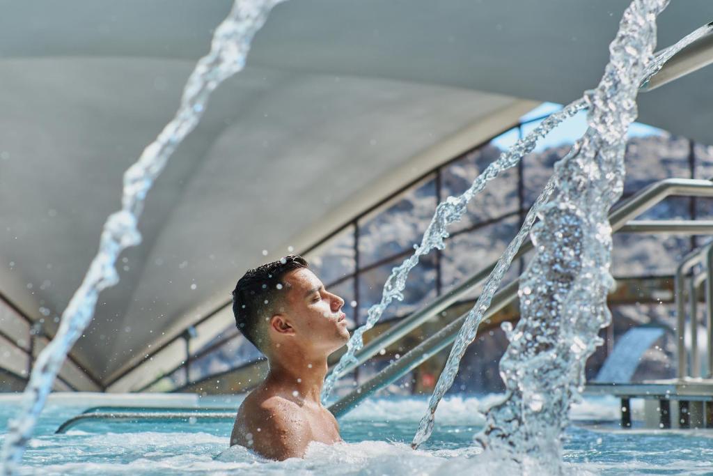 Jaunais vīrietis relaksējas ūdenī, kamēr no strūklakas plūst ūdens. Skats uz modernu arhitektūru un kalnu fona.