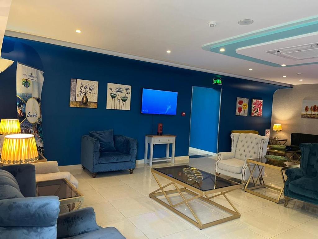 Mūsdienīgs viesnīcas interjers ar zilu sienu, elegantiem dīvāniem un zelta akcentiem. Telpā ir televizors un mākslas darbi uz sienām, radot viesmīlīgu atmosfēru.