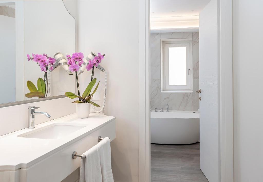 Mūsdienīga vannas istaba ar baltu izskatu, kurā ir eleganta izlietne un purpura orhidejas pušķis uz galda. Aiz izlietnes redzama caurspīdīga durvju atvēršana uz vannu ar logu.