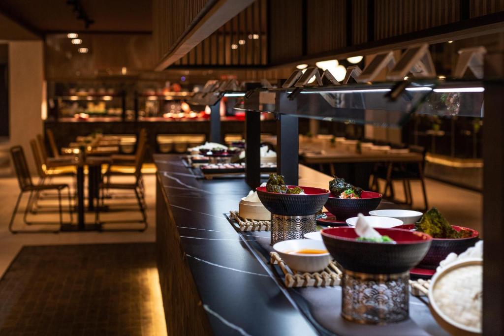 Elegants restorāns ar modernu interjeru, kurā redzami dažādi ēdieni uz bufetes galda. Apgaismojums rada patīkamu atmosfēru, un fonā ir redzami galdi un krēsli.