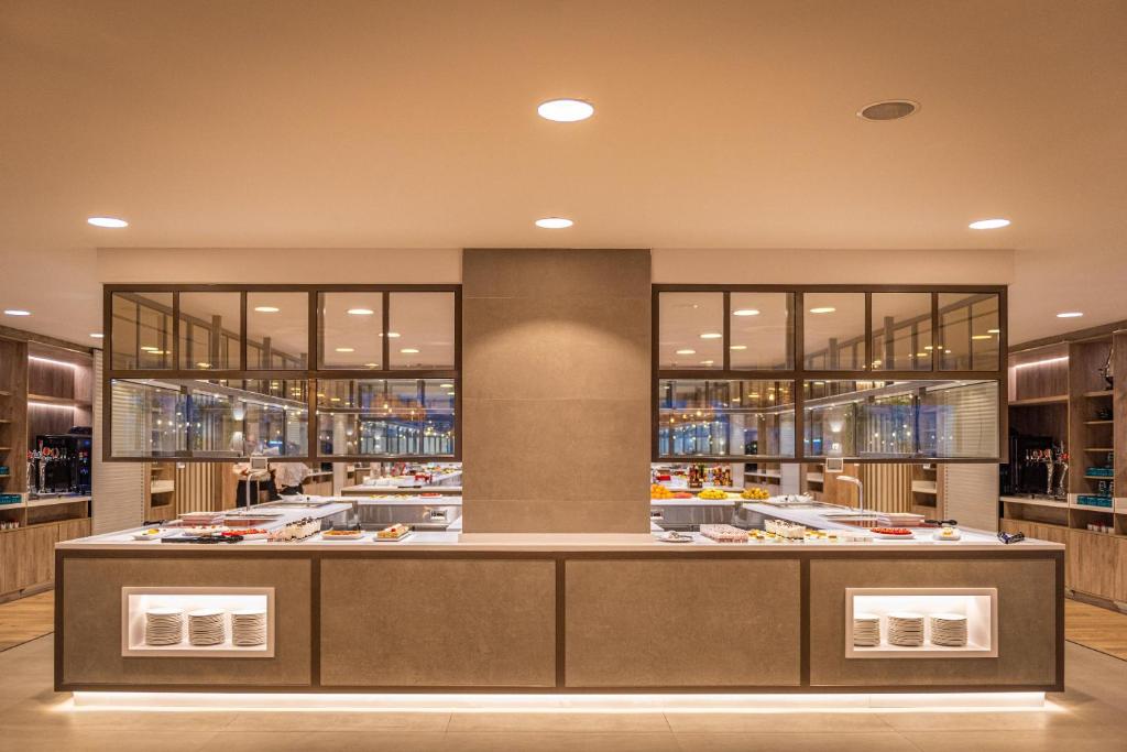 Mūsdienīga restorāna iekārtojums ar elegantu bufeti, kurā redzami dažādi ēdieni un dzērieni. Apgaismojums rada siltu un aicinošu atmosfēru.