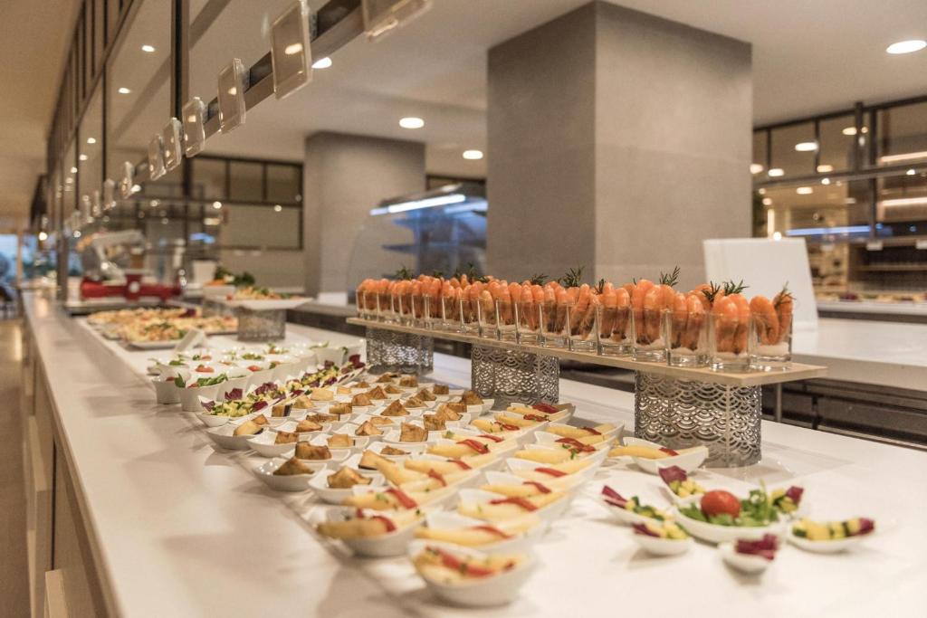 Piedāvājums bufetē ar dažādiem ēdieniem, tostarp garnelēm un maziem uzkodu traukiem, kas izkārtoti uz baltas virsmas.