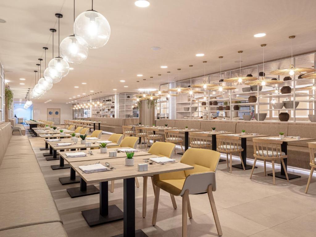 Mūsdienīgs restorāns ar gaišu interjeru, elegantiem galdiem un krēsliem, kas rotāti ar zaļiem augiem. Plakanie gaismekļi un stilīgas plauktu sistēmas rada patīkamu atmosfēru.