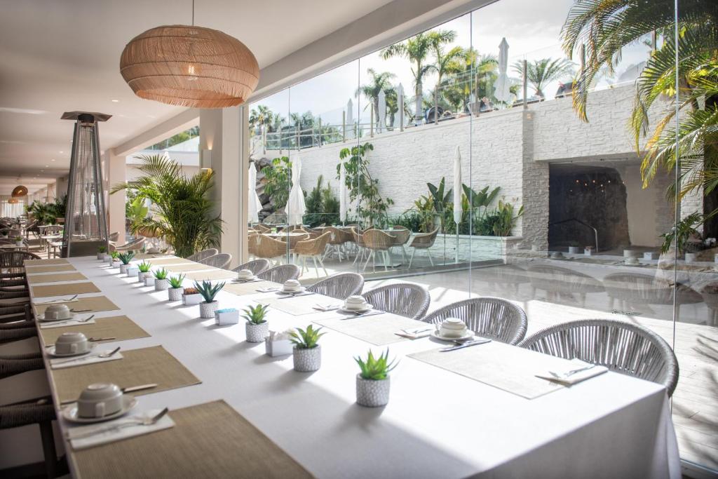 Mūsdienīgs restorāna interjers ar garu galdu, dekorētu ar sulīgām augiem un traukiem, caur plašiem logiem redzama zaļa iekārtojuma zona.