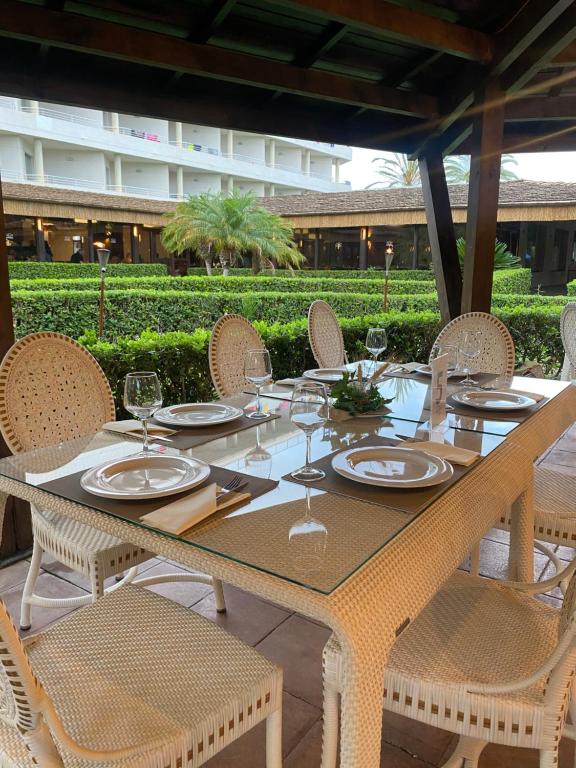 Elegants galda iestatījums ar stikla virsmu un rotājumu, kurā redzami balti trauki un glāzes, atrodas zem jumta ar koka balstiem, apkārt ir zaļi krūmi un palma.