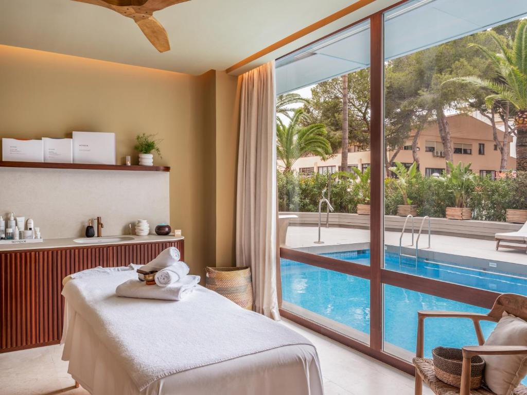 Mūsdienīgs spa telpas interjers ar masāžas galdu, kosmētikas produktiem un lieliem logiem, kas paver skatu uz baseinu un tropisko dārzu.