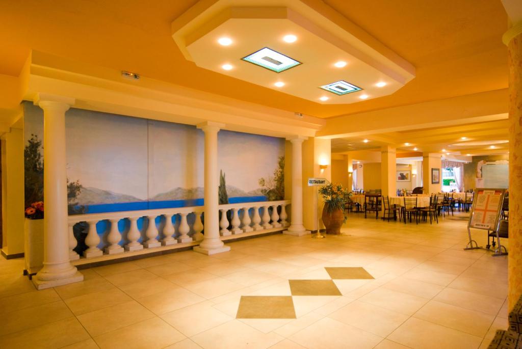 Restorāna interjers ar elegantu balto kolonnu dizainu un gleznu uz sienas, kas attēlo jūras skatu. Telpa ir gaiša un plaša, ar koka galdiem un krēsliem.
