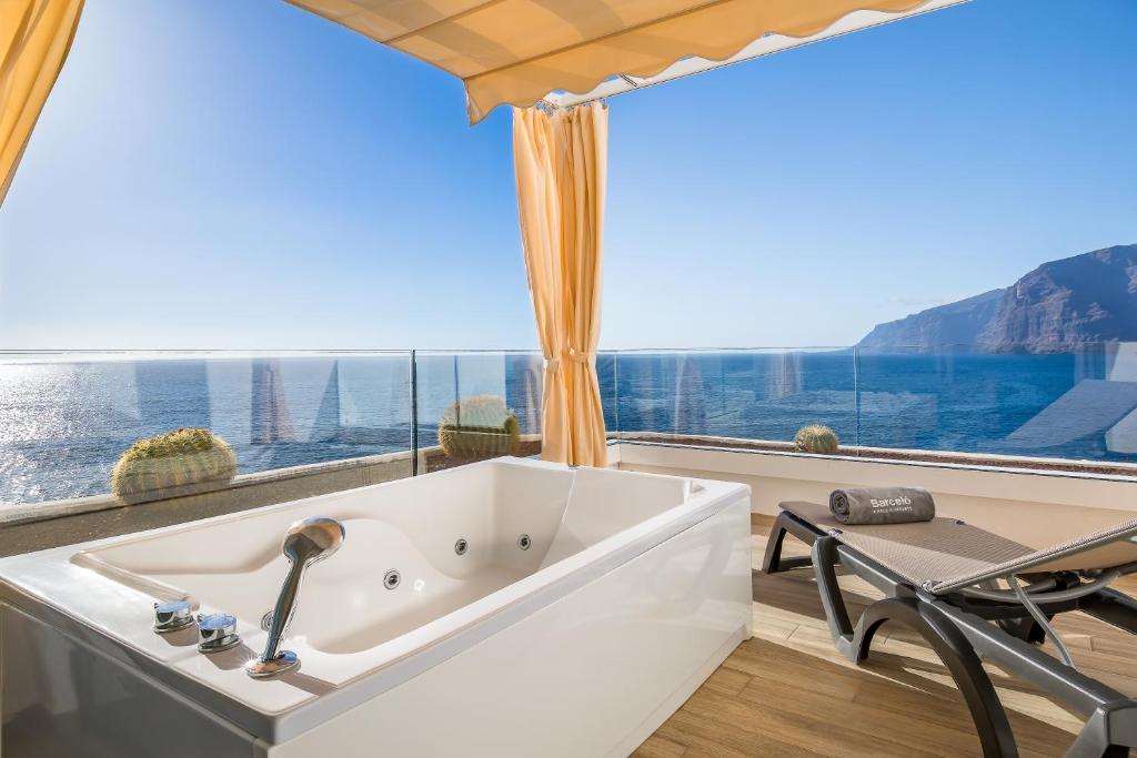 Luksusa terase ar jacuzzi un skatu uz jūru, kurā ir saules krēsli un dzeltenas aizkari. Ideāla vieta relaksācijai un atpūtai.