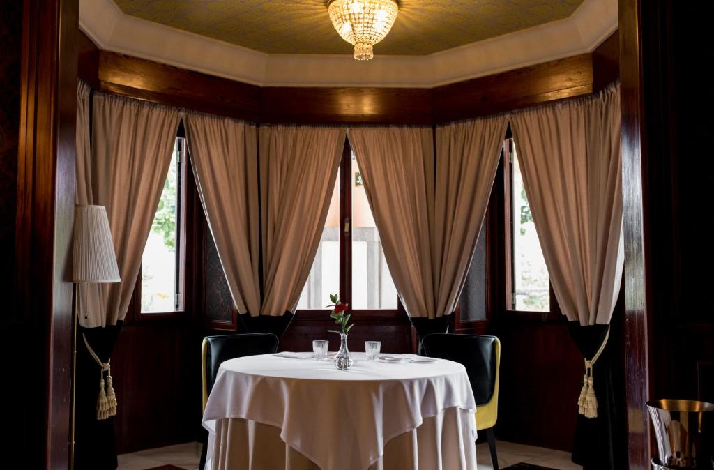 Eleganta restorāna iekšiene ar apaļu galdu, baltu galdautu un modernu apgaismojumu. Logi ar aizkariem rada intīmu atmosfēru, ideālu romantiskām vakariņām.