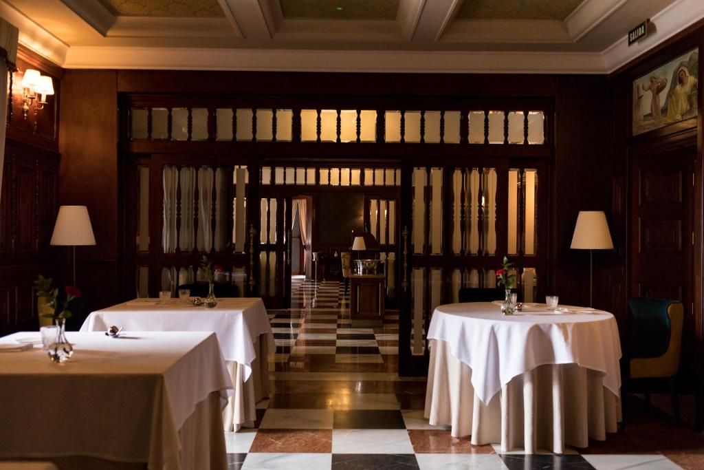 Elegants restorāna interjers ar baltiem galdautiem un ziediem uz galdiem, kas veido izsmalcinātu atmosfēru. Gaisma maigi apgaismo koka detaļas un eleganto grīdu.