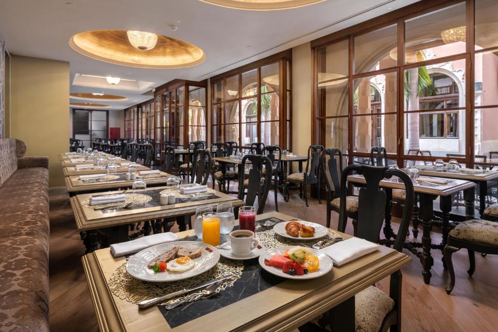 Eleganti iekārtota brokastu telpa ar galdiem, kas klāti ar dažādiem ēdieniem, tostarp augļiem un maizēm. Labi apgaismota telpa ar lieliem logiem, kas sniedz skatu uz ārpusi.