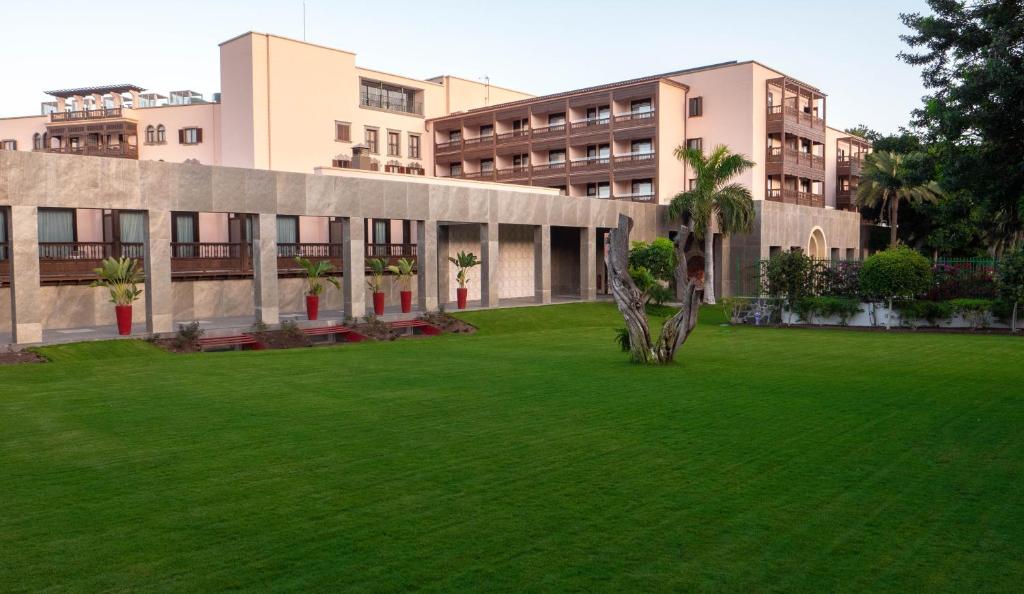 Zaļš zāliena laukums ar modernu arhitektūru un dekoratīvām palmu stādām, kas atrodas viesnīcas teritorijā. Fons veido elegance un mierīgu atmosfēru.