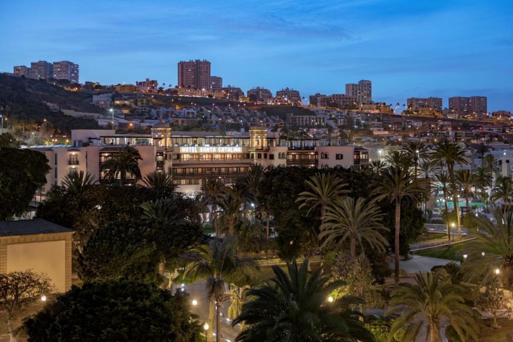 Pilsētas ainava ar viesnīcu "Hotel Santa Catalina" un palmu stādījumiem, apgaismota vakara stundā. Fons veido modernu apbūvi kalnā.