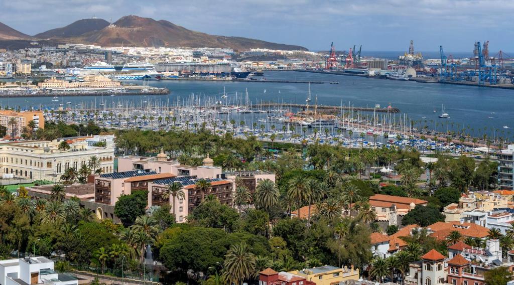 Skats uz Laspalmas ostu, kur redzami jahtu piestātne, kravas kuģi un kalnu foni. Apstādījumi un pilsētas arhitektūra veido dzīvīgu ainavu.