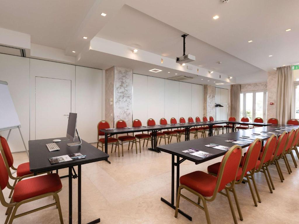 Konferenču zāle ar galdiem un sarkaniem krēsliem, aprīkota ar projektoru un prezentācijas iekārtām. Telpa ir gaiša un moderna, ideāli piemērota sanāksmēm un semināriem.