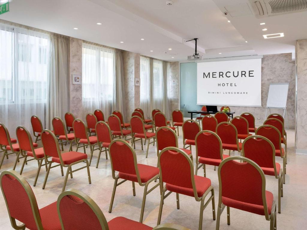 Konferenču telpa Mercure viesnīcā Riminī, ar sarkaniem krēsliem un projektoru, gatava pasākumam.