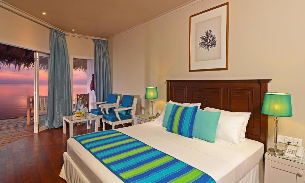 Mūsdienīga viesnīcas numura interjers ar gultu, spilveniem un krēsliem, kas piedāvā skatu uz jūru. Logi atveras uz terasi, kur ir iespējams baudīt saulrietu.