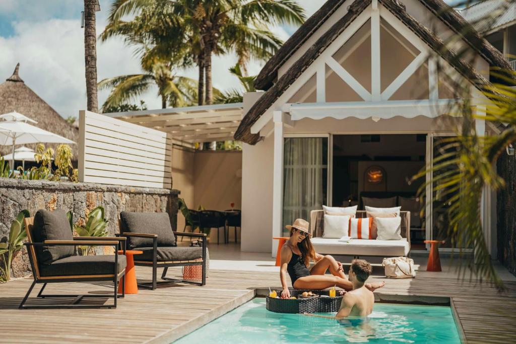 Atpūtas brīvdienu ainava ar baseinu, kurā divi cilvēki relaksējas, baudot sauli un dzērienus. Apkārt ir tropiska daba un mūsdienīga māja ar terasi.