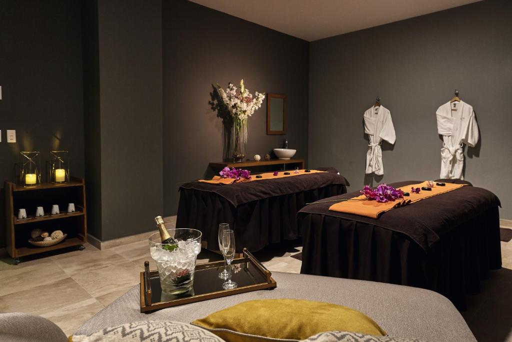 Relaksējoša spa telpa ar masāžas galdiem, uz kuriem novietoti ziedi un dvieļi. Blakus atrodas džakuzi ar šampanieti un glāzēm, kā arī baltas halāti. Telpā ir mierīga atmosfēra ar ziediem un svecēm.