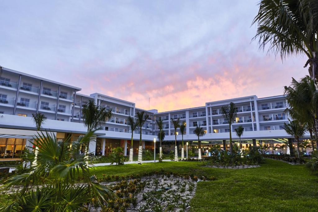 Mūsdienīgs viesnīcas komplekss ar zaļu apstādījumu un palmām, kas atrodas saulrietā. Gaismas elementi piešķir atmosfēru, radot relaksējošu vidi.