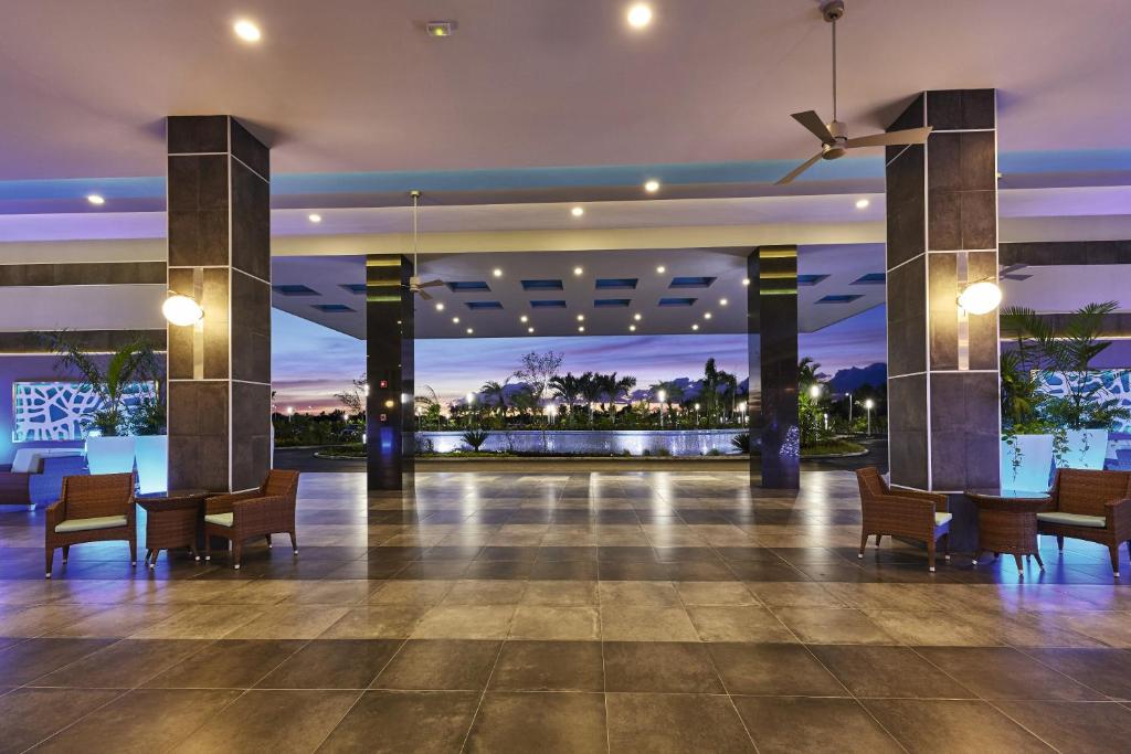 Mūsdienīgs viesnīcas vestibils ar elegantām mēbelēm un apgaismojumu, kas rada relaksējošu atmosfēru. Aiz logiem redzama ainava ar zaļumiem un ūdens elementiem.