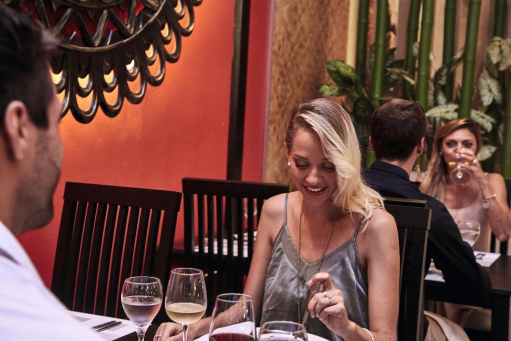 Eleganta vakariņu aina restorānā, kur divi pāri izbauda maltīti un sarunas, kamēr viens no viesiem smaidot skatās uz savu šķīvi. Siltas krāsas un zaļumi piešķir atmosfērai patīkamu noskaņu.