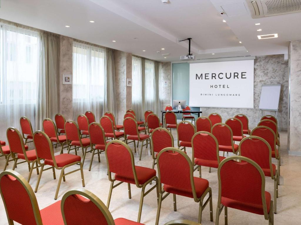Konference telpa Mercure Hotel, kurā ir sarkanas krēsli un projekcijas ekrāns. Telpa ir gaiša un mūsdienīga, ideāla sanāksmēm un prezentācijām.
